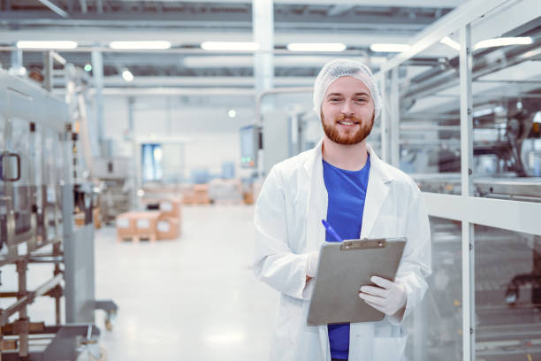 joven guapo sonriente científico con portapapeles posando en fábrica - industry portrait production line factory fotografías e imágenes de stock