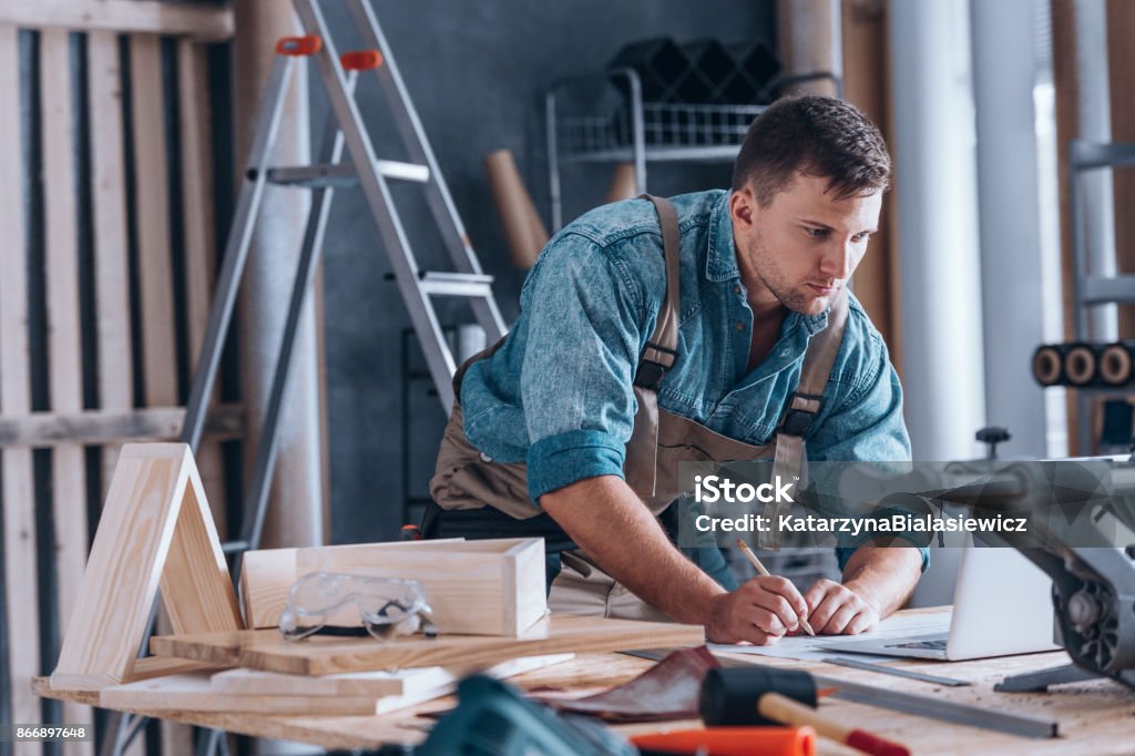 Handsome carpenter working in workshop Handsome carpenter working on project at table with tools and crafts in his workshop using laptop and pencil Small Business Stock Photo