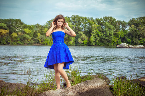 Portrait of beautiful young brunette woman, wearing elegant blue dress, standing on a stone river bank with green trees and blue sky on background