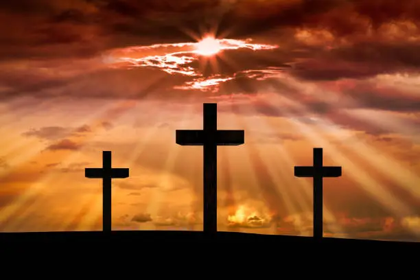 Jesus Christ cross on a scene with dramatic sky, lighting, red, orange sunset, clouds, sunbeams, sun rays glowing behind three crosses on Golgotha mountain. Easter, resurrection, Good Friday, Calvary concept.