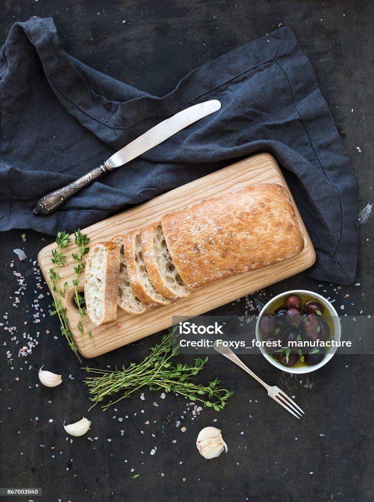 Italian ciabatta bread cut in slices Italian ciabatta bread cut in slices on wooden chopping board with herbs, garlic and olives over dark grunge backdrop, top view Backgrounds Stock Photo