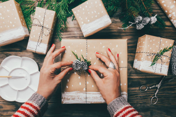 Woman wrapping Christmas presents in a crafty way Woman´s hands wrapping Christmas presents on brown paper decorated with painted snow, fir branches and pinecones on a rustic wooden board. Top view wrapping paper stock pictures, royalty-free photos & images