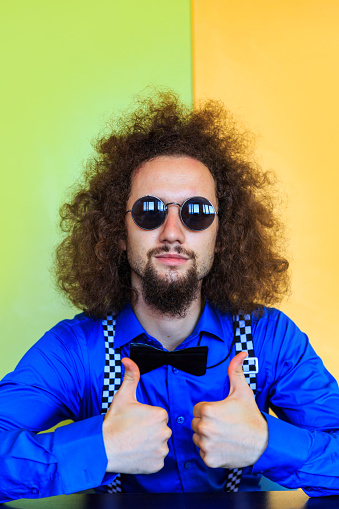 Portrait of young man with sunglasses, blue shirt, bowtie and suspenders in front of colored background.
