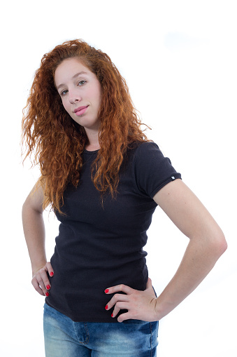 Young woman is holding her hand on her waist, she looks confidently. The person is redheaded and young. White background. Black shirt.