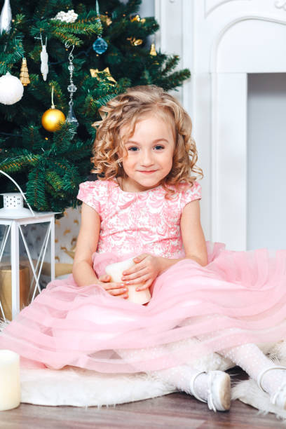 słodka blondynka z lokami w różowej sukience siedzi w dekoracjach noworocznych w pobliżu drzewa ozdobionego zabawkami - flower gift decoration domestic room zdjęcia i obrazy z banku zdjęć