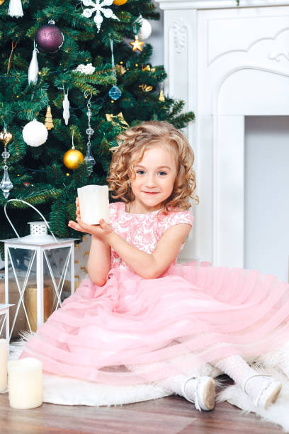 małe dziecko siedzi pod choinką w pięknej różowej sukience, trzyma świecę, wokół prezentów - flower gift decoration domestic room zdjęcia i obrazy z banku zdjęć