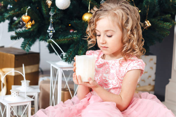 blond dziewczyna w różowej sukience pod drzewem noworocznym i patrzy na płonącą świecę - flower gift decoration domestic room zdjęcia i obrazy z banku zdjęć