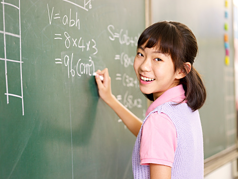 asian elementary schoolgirl looking at camera smiling while solving a math problem on chalk board.