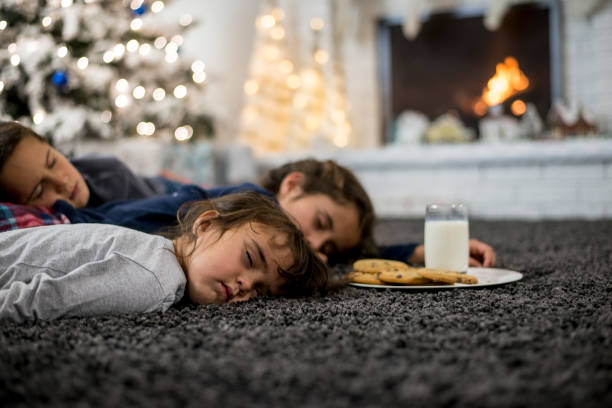 Three siblings are indoors in their living room during Christmas. They are wearing warm, comfortable clothes. They have fallen asleep while waiting for Santa with a glass of milk and cookies.