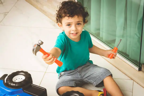 Cute little boy, sitting on the floor, and showing a toy hammer and a toy screwdriver.