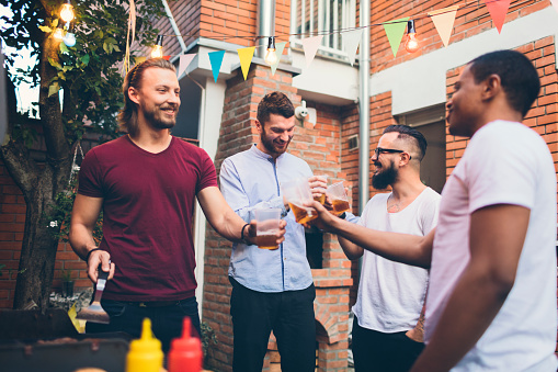 Male friends having fun on a barbecue in back yard. Standing by the gas grill and grilling meat. Talking and drinking beer, alcohol from transparent disposable cups and cheering.