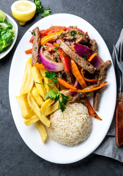 Peruvian dish Lomo saltado - beef tenderloin with purple onion, yellow chili, tomatoes served on white plate with french fries and rice. Top view