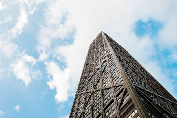 This is a horizontal, color photograph of the iconic Hancock Building in downtown Chicago, Illinois. Photographed from a low angle view. The sky is blue with white clouds.