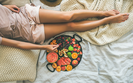 Summer healthy raw vegan clean eating breakfast in bed concept. Young girl wearing pastel colored home clothes taking fruit from tray full of fresh seasonal fruit. Top view, copy space