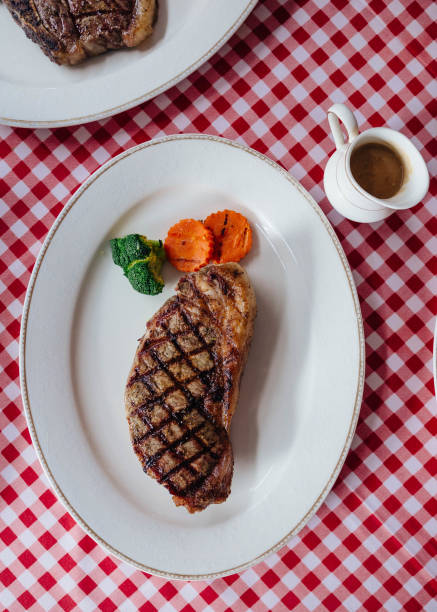 vista dall'alto del wagyu disossato alla griglia charcoal top loin bistecca servita con salsa barbecue e patate al forno in piatto bianco su tovaglia a motivi rossi e bianchi. - steak strip steak prepared potato grilled foto e immagini stock