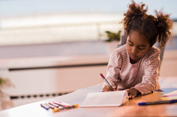 istock Little black girl writing in notebook while relaxing on the table. 867333432
