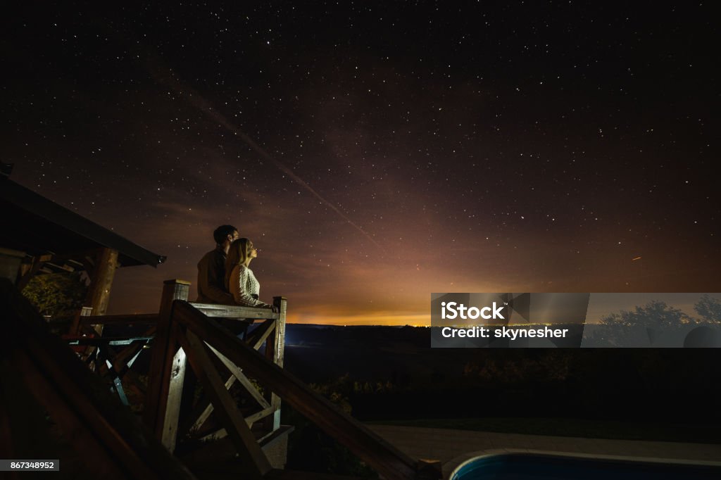 Loving couple looking at breathtaking view of constellation from their balcony. Young couple enjoying in a romantic view on constellation from their terrace by night. Copy space. Night Stock Photo