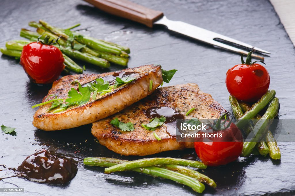 Grilled pork chops, steaks with vegetables, tomatoes, beans and sauce on a black slate. Fresh meat with foam. Dark background. Grilled and barbecue concept. Backgrounds Stock Photo