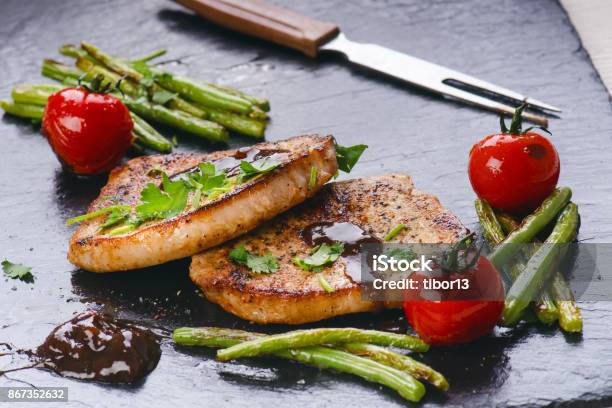 Grilled Pork Chops Steaks With Vegetables Tomatoes Beans And Sauce On A Black Slate Fresh Meat With Foam Dark Background Grilled And Barbecue Concept Stock Photo - Download Image Now