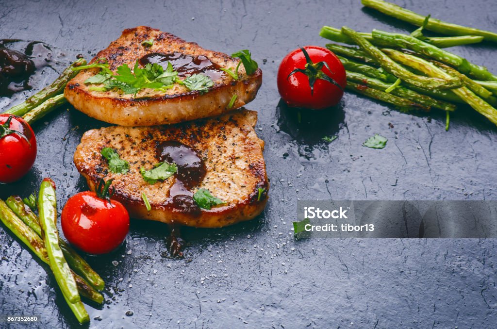 Grilled pork chops, steaks with vegetables, tomatoes, beans and sauce on a black slate. Fresh meat with foam. Dark background. Grilled and barbecue concept. Backgrounds Stock Photo