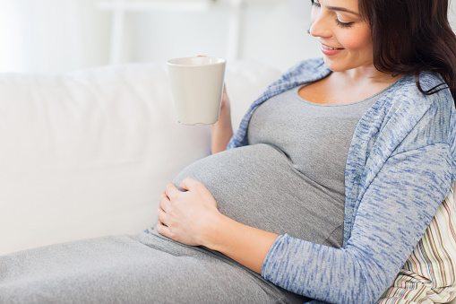 pregnancy, drinks, rest, people and expectation concept - close up of happy pregnant woman with big belly holding cup and drinking tea at home