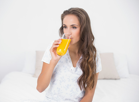 Calm attractive brunette drinking a glass of orange juice in bright bedroom