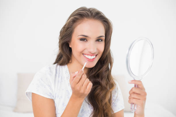 istock Smiling attractive brunette applying lip gloss and holding mirror 867435700