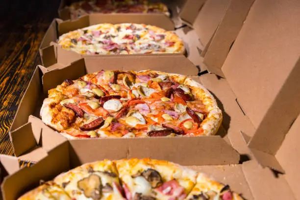 Close up of tasty pizzas with variety of toppings and cheese in cardboard take out boxes with open lid on wooden table