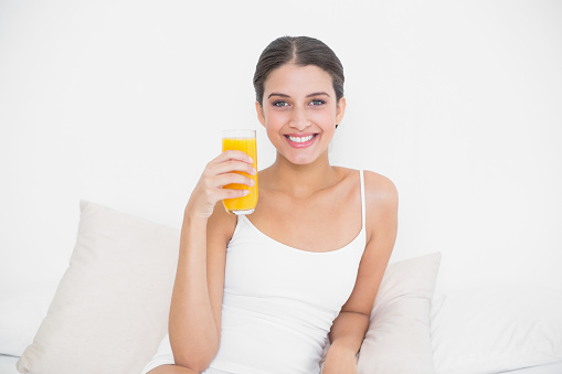 Dynamic young brown haired model in white pajamas holding a glass of orange juice in bright bedroom