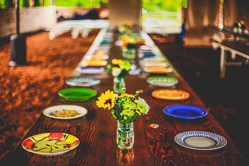 Looking down a long, rustic, wooden dining table set with multi-colored, multi patterned yellow and blue plates and mason jar vases filled with sunflower and wildflower bouquets