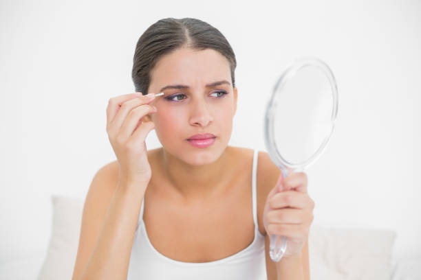 istock Frowning young brown haired model in white pajamas plucking her eyebrows 867528374