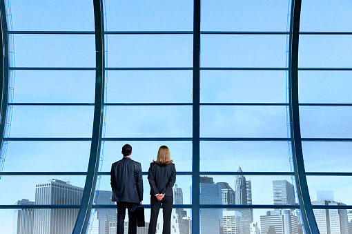 A rearview of a businessman and a businesswoman standing side by side as they look out a large office building window towards a skyline in the distance.