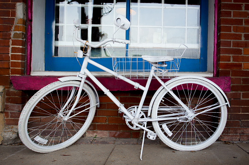 An old pedal bike painted white leaning on a kickstand in front of a red brick wall and multi paned window