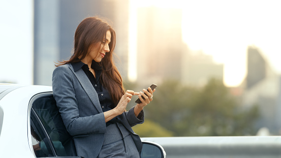 Business Woman Uses Smartphone While Leaning on Her Premium Class Car. Big City with Skyscrapers in the Background.