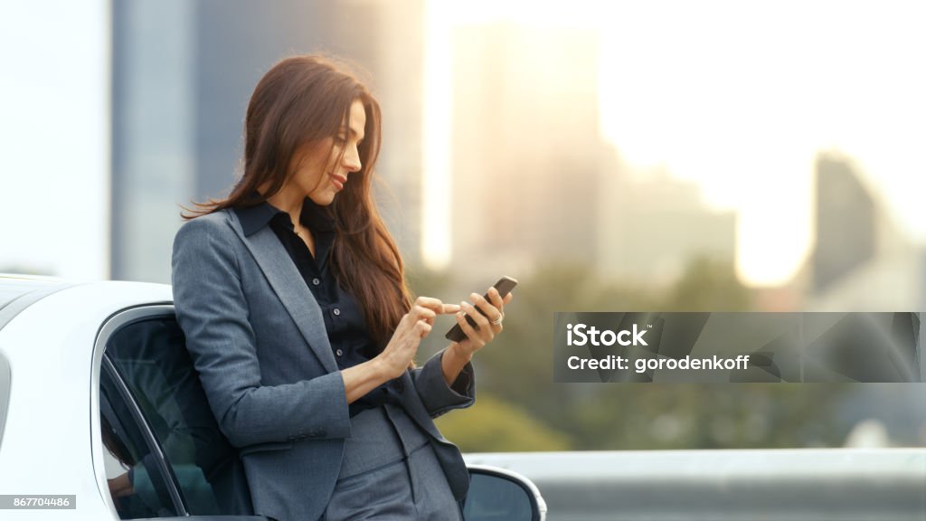 Business Woman Uses Smartphone While Leaning on Her Premium Class Car. Big City with Skyscrapers in the Background. Car Stock Photo