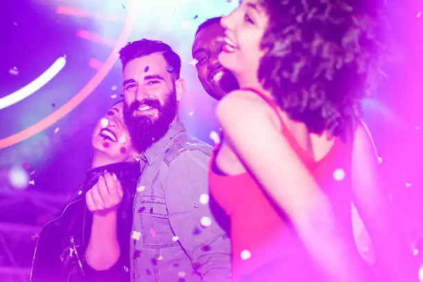 Photo of Happy friends having fun in night club with canon ball throwing confetti - Young people enjoying weekend nightlife with original laser lights color - Soft focus on bearded white man - Warm filter