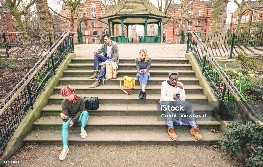 Group of multiracial friends using mobile smart phone sitting on stairs at park - Young hipster people addicted by smartphone on social network community - Technology concept - Desaturated color tones Mobile App Stock Photo