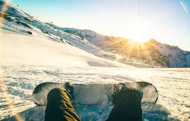 Snowboarder sitting at sunset on relax moment in french alps ski resort - Winter sport concept with adventure guy on top of mountain ready to ride down - Legs view point with teal and orange filter