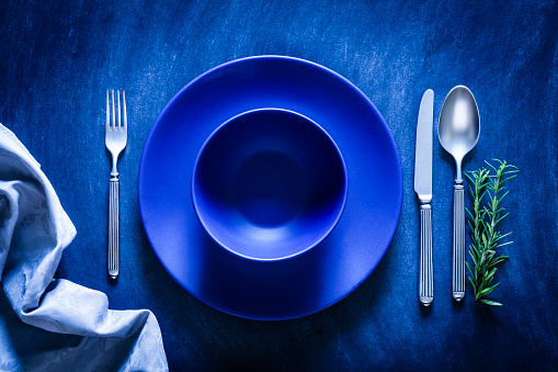 Top view of a blue plate and bowl with a spoon, fork and knife at each side shot against a textured bluish toned background. A gray textile napkin is at the bottom-left corner of an horizontal frame. A rosemary twig is also beside the plate. Predominant color is blue. Low key DSRL studio photo taken with Canon EOS 5D Mk II and Canon EF 100mm f/2.8L Macro IS USM
