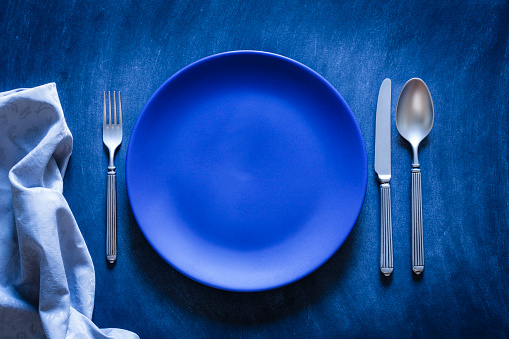 Top view of a blue plate with a spoon, fork and knife at each side shot against a textured bluish toned background. A gray textile napkin is at the bottom-left corner of an horizontal frame. Predominant color is blue. Low key DSRL studio photo taken with Canon EOS 5D Mk II and Canon EF 100mm f/2.8L Macro IS USM