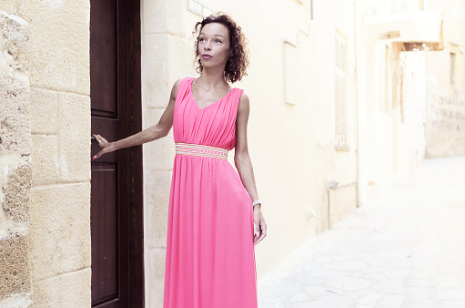 Slim sexy woman in long pink dress holding wooden door and walking in narrow street.