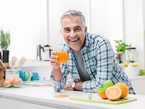 Smiling man having breakfast at home, he is preparing and drinking a glass of fresh orange juice, kitchen interior on the background