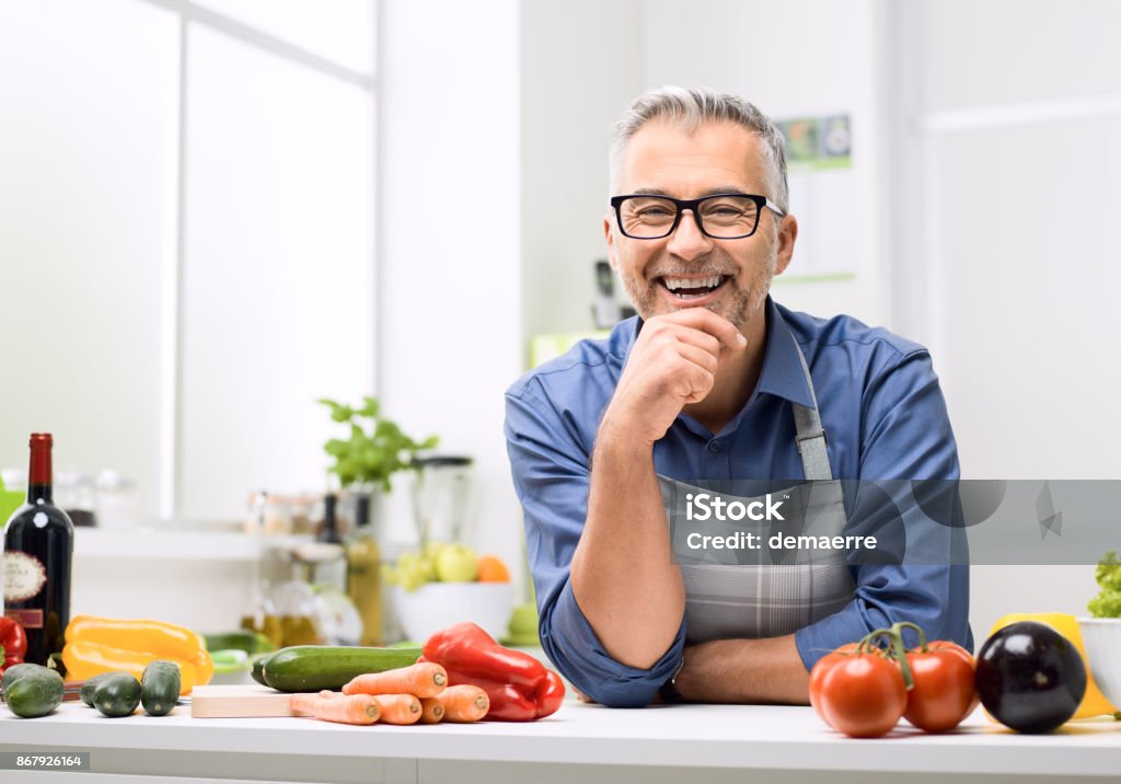 Confident smiling man posing in his kitchen Smiling man posing in the kitchen, he is preparing healthy homemade food using fresh organic vegetables Men Stock Photo