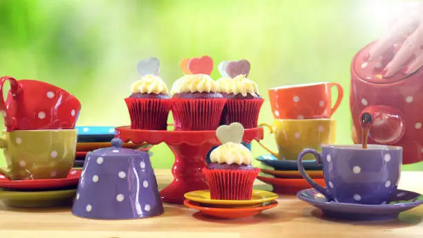 Colorful Mad Hatter style tea party with cupcakes and rainbow colored polka dot cups and saucers, with bokeh garden background and lens flare, pouring tea.