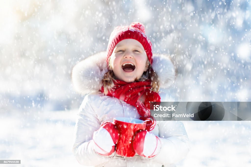 Child drinking hot chocolate in winter park. Kids in snow on Christmas. Child drinking hot chocolate with marshmallows in snowy winter park. Kid with cup of warm cocoa drink on Christmas vacation. Little girl playing in snow on Xmas eve. Family outdoor winter fun. Child Stock Photo