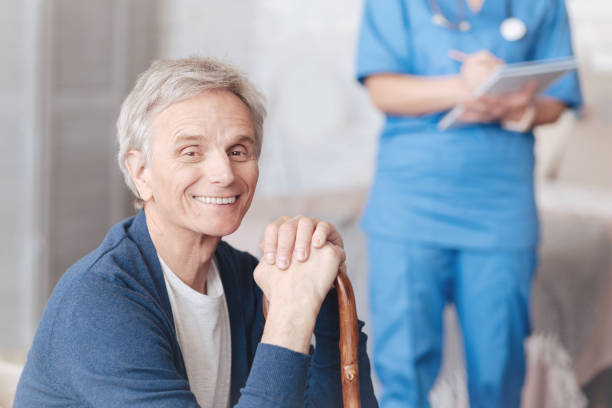 Radiant senior man smiling cheerfully into camera Positivity is healing. Positive minded elderly gentleman leaning on his walking cane and looking into the camera with a cheerful smile on his face while a medical worker taking notes aids walk stock pictures, royalty-free photos & images