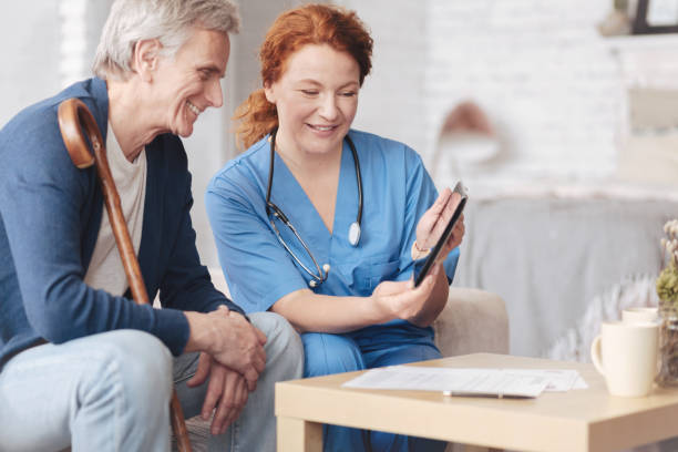 Friendly senior patient and nurse looking tablet computer Positive thinking. Side view on a positive minded elderly man and a female doctor smiling while sitting on a sofa and looking at a screen of a digital tablet of home. aids walk stock pictures, royalty-free photos & images