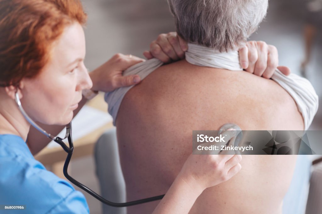 Close up of doctor listening back of patient with stethoscope Regular check up. Scaled up look on serious female medical worker focusing her attention while holding a stethoscope and listening to lung sounds of a male patient. Lung Stock Photo