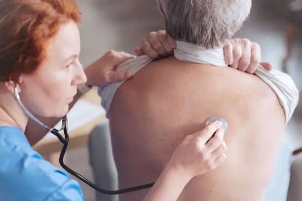 Regular check up. Scaled up look on serious female medical worker focusing her attention while holding a stethoscope and listening to lung sounds of a male patient.