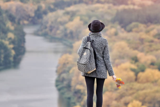 istock Girl with a backpack and a hat standing on a hill. River and forest below. Back view 868038286